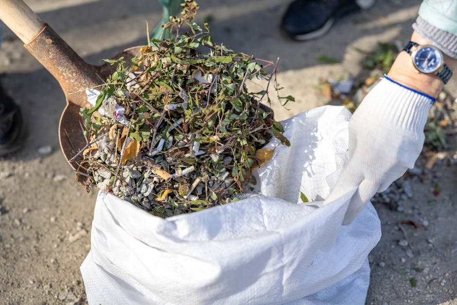 A person wearing a white gardening glove is holding a white sack filled with garden waste, including trimmed ivy, small branches, and gravel. The individual is using a trowel or small shovel with a wo